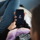 A young woman sits back on the couch holding a phone that has the TikTok logo on it.
